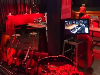 Színpad vonós hangszerekkel és egy zongoristát mutató képernyővel, vörös fényben