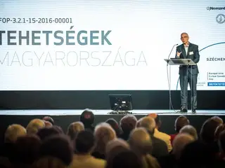Előadó a pulpitusnál a Tehetségek Magyarországa felirat előtt, előtérben a közönség